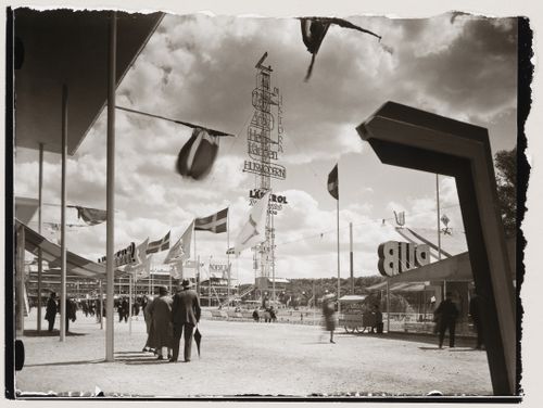 Exterior view of the advertising mast and Paradise Restaurant at the Stockholm Exhibition of 1930 showing a walkway and flagpoles, Stockholm