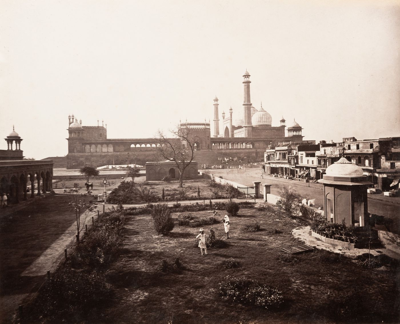 View of the Jami Masjid from the south showing commercial buildings on the right and a garden in the foreground, Delhi (now Delhi Union Territory), India