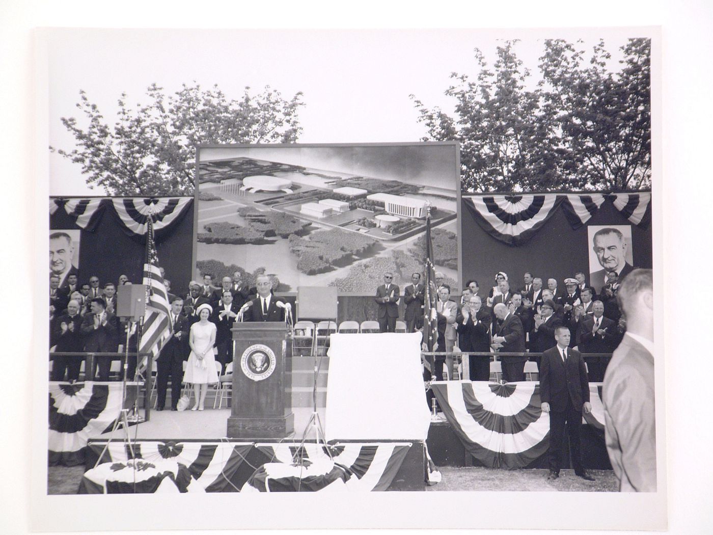 View of the dedication ceremony of the John F. Kennedy Educational, Civic and Cultural Center showing a partial view of the platform with people, posters of President Lyndon B. Johnson and a photograph of the model of the building, Mineola, New York