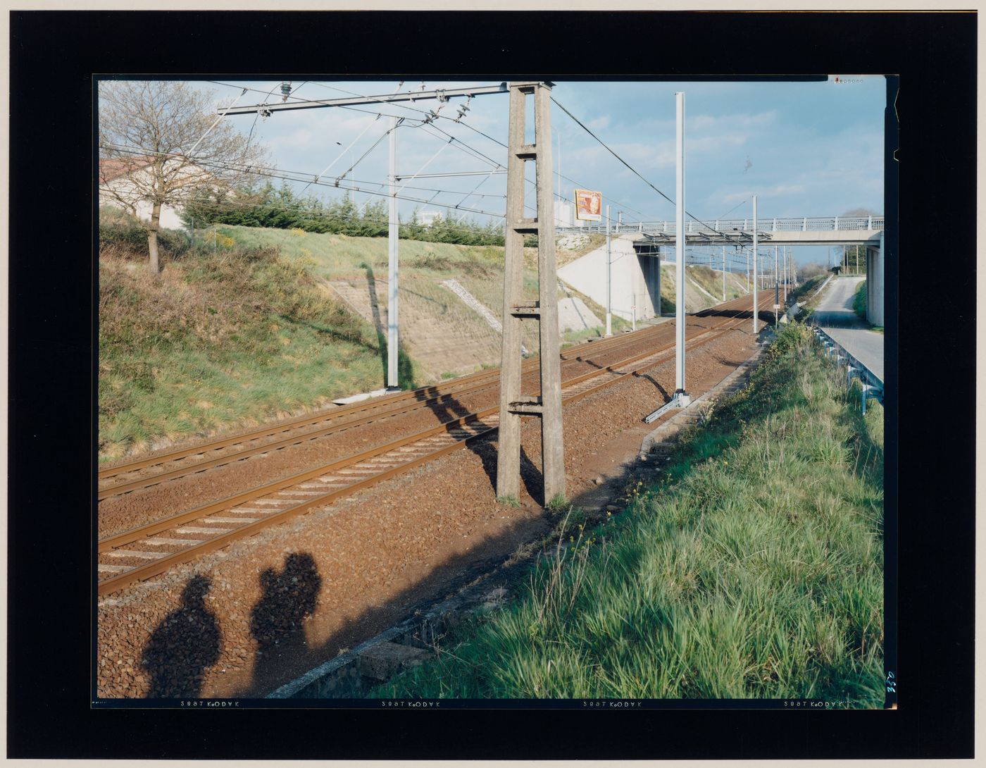 View of tracks and overhead wires for an electric railway, a grassy slope, a bridge and the cast shadows of the photographer and another person, Orthez, France (from the series "In between cities")