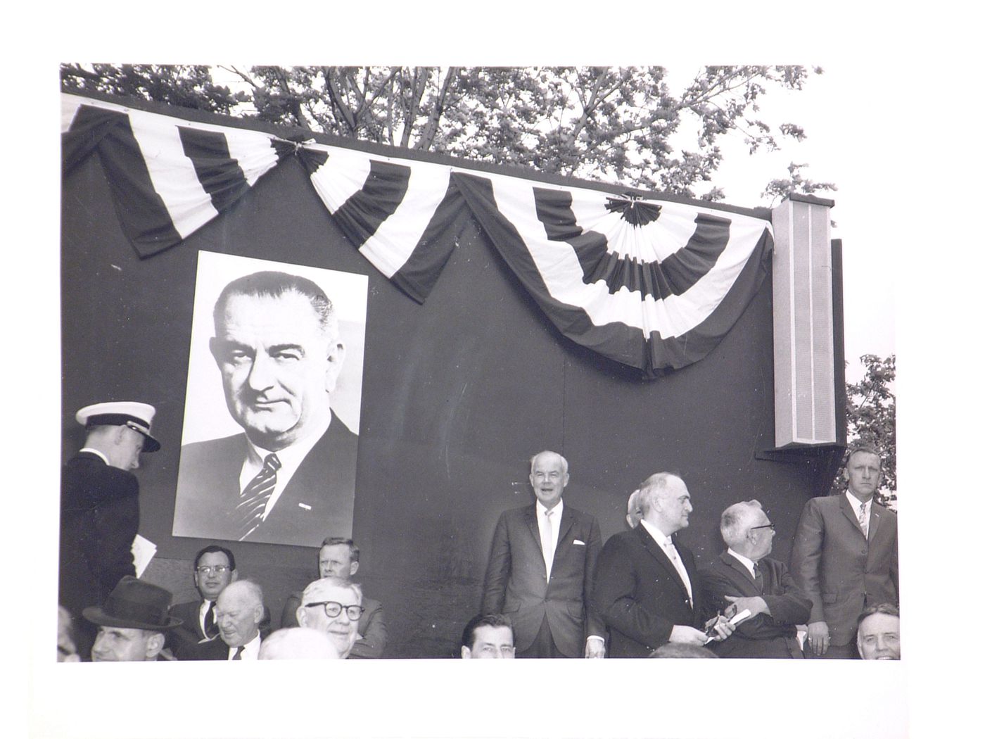 View of the dedication ceremony of the John F. Kennedy Educational, Civic and Cultural Center showing a close-up view of a poster of President Lyndon B. Johnson, a photograph of the model of the building and people on the platform, Mineola, New York, Unit