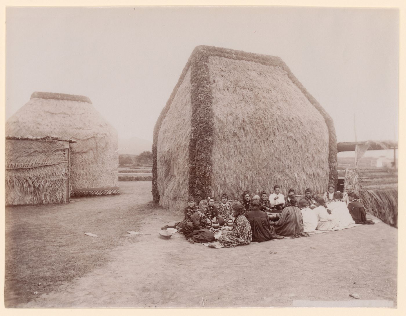 View of two "hale pili" (grass-covered buildings), with group of people at a lū'au in the foreground, Moanalua Gardens, Honolulu, Hawaii, United States