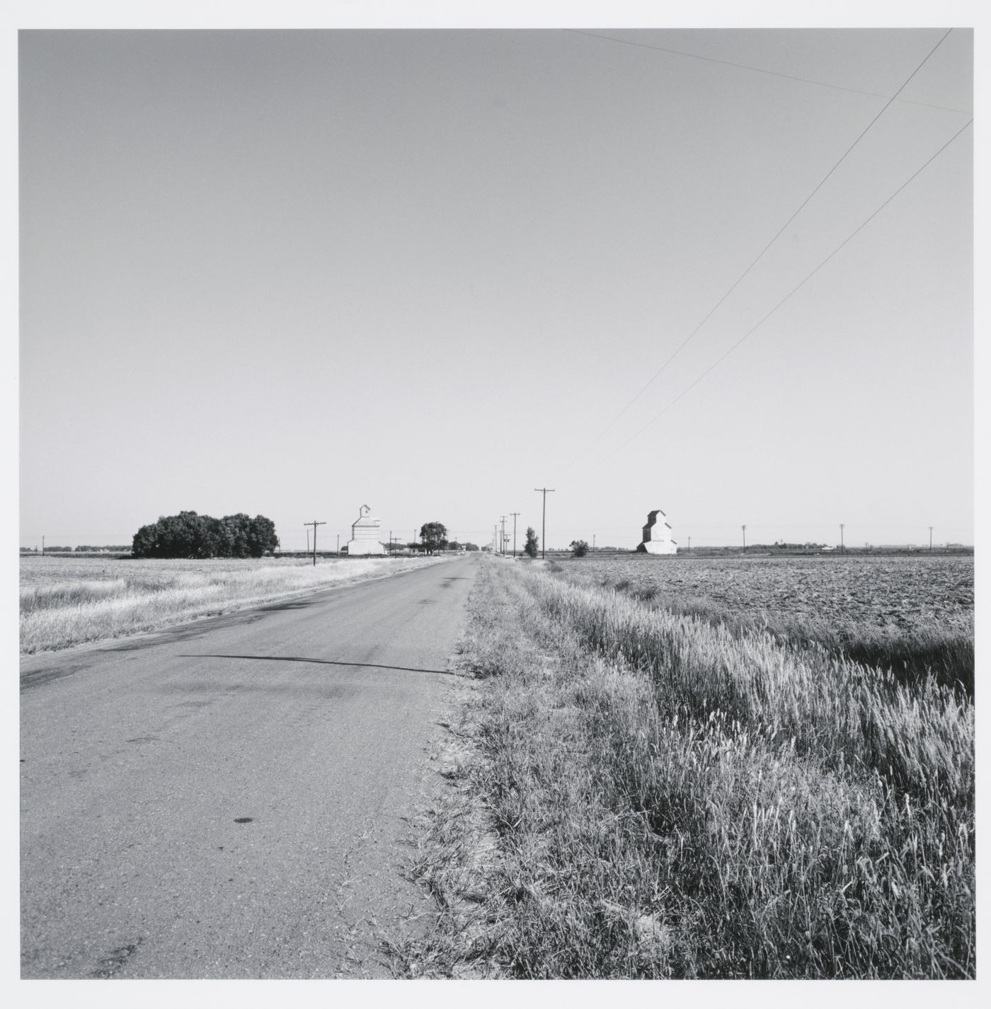 View of road with fields and two grain elevators on either side, Kinsley, Kansas