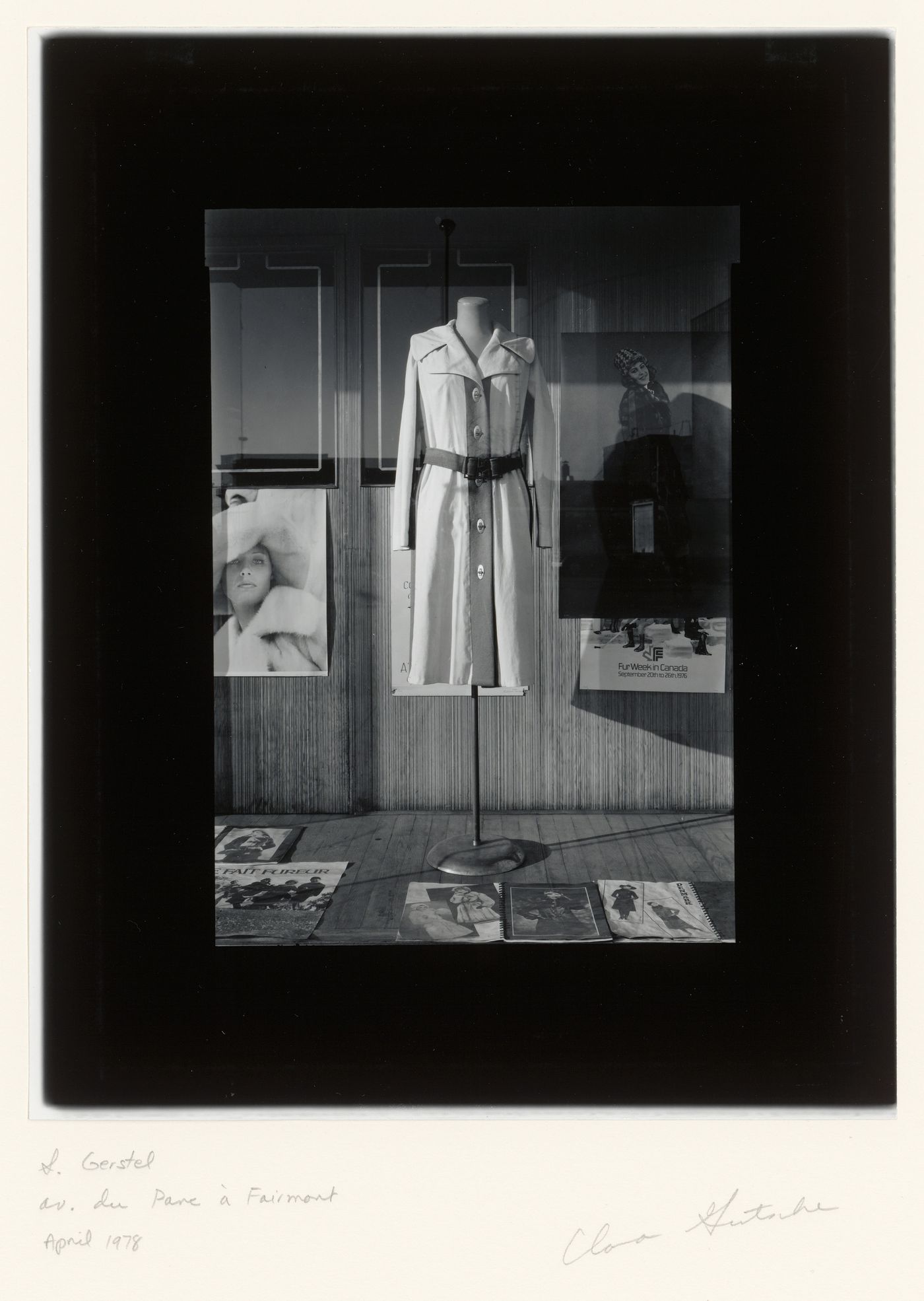 View of a display window of the S. Gerstel clothing store showing an overcoat, fashion catalogues and posters and the reflection of buildings across the street, corner of avenue du Parc and rue Fairmount Ouest, Montréal, Québec