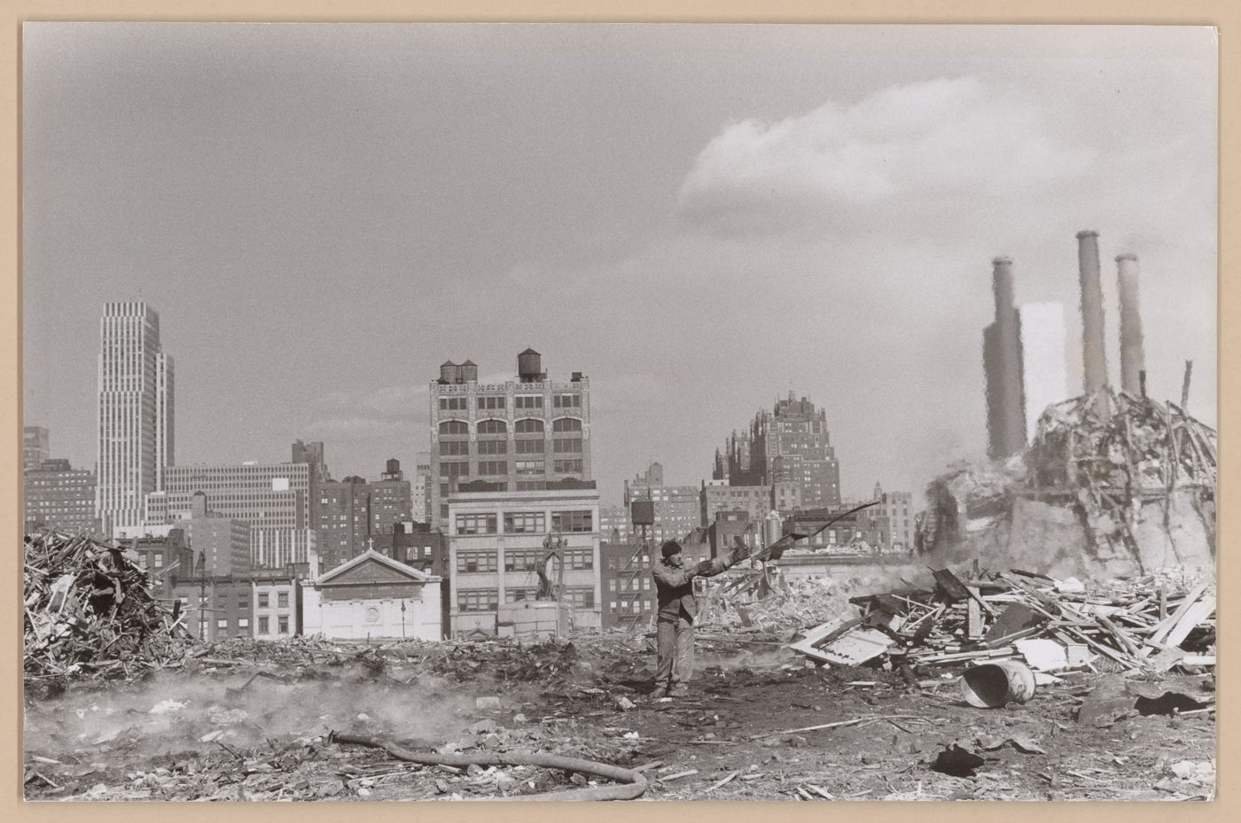 View of a demolition site [?] showing a man and fire hose with skyscrapers and smokestacks in the background, New York City, New York