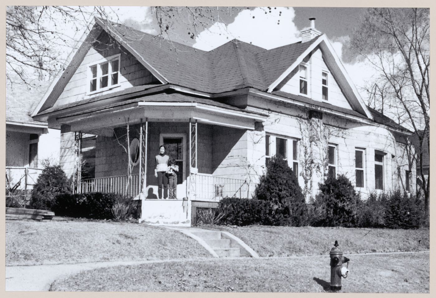 Photograph showing an exterior view of the house used for Clay House prior to being covered in clay along with an adult and a child standing in the house's doorway