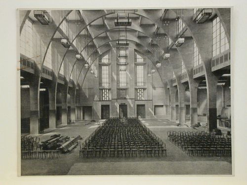 Interior view of a building hall, possibly a church, showing arches, windows and aligned chairs