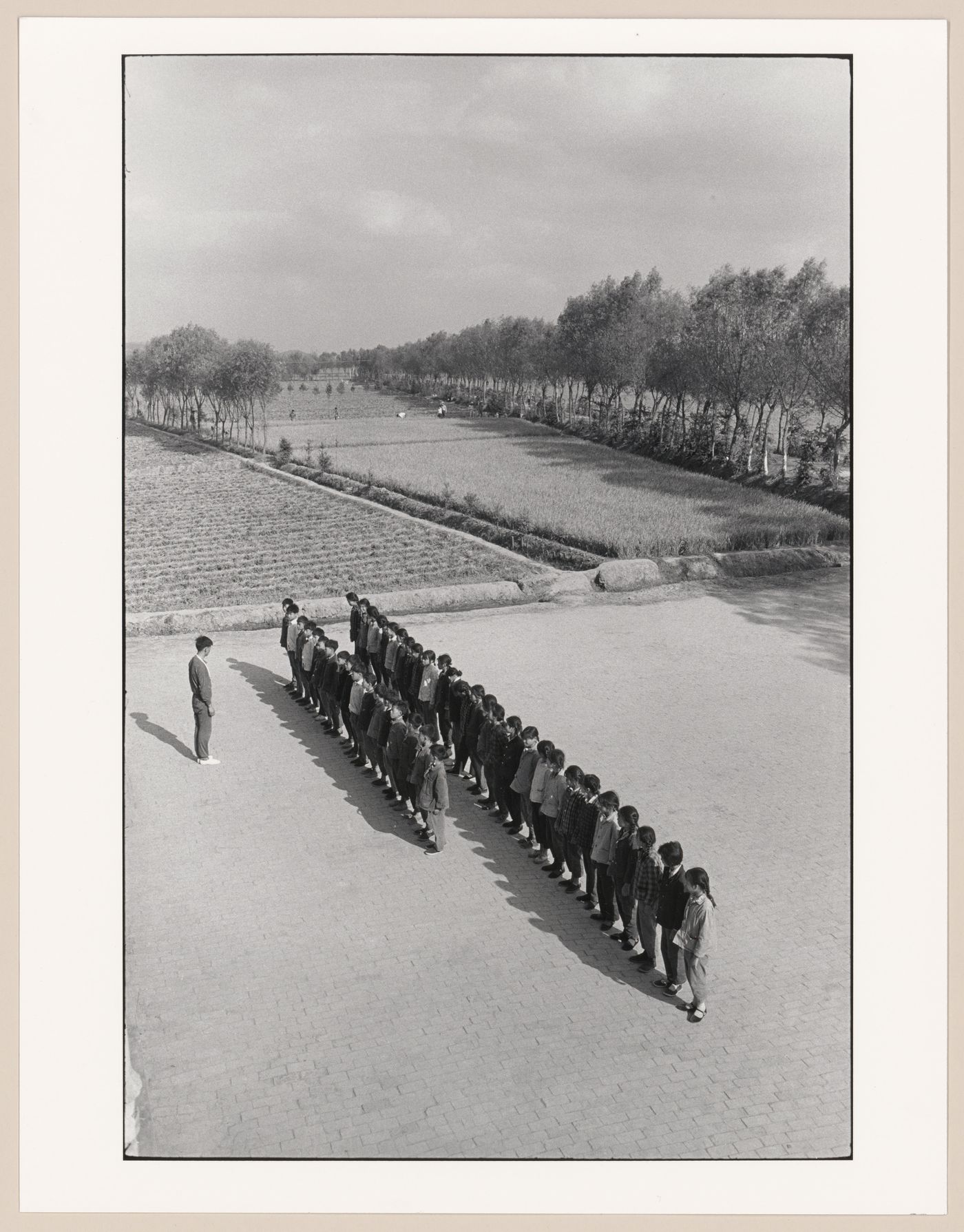 Partial view of a collective farm, with a teacher and students exercising in the foreground and workers harvesting rice in the background, Hua Xi, Jiangsu Sheng, China