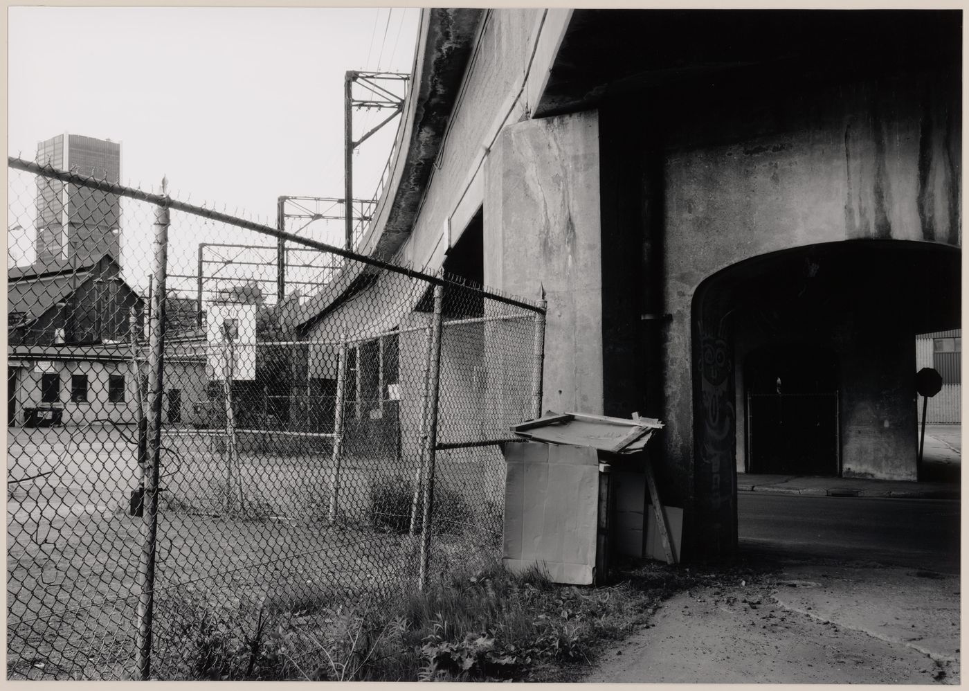 Field Work in Montreal: View of a wood and cardboard shack-like structure, a chain link fence, a railroad bridge and graffiti showing an office building in the background, Montréal, Québec