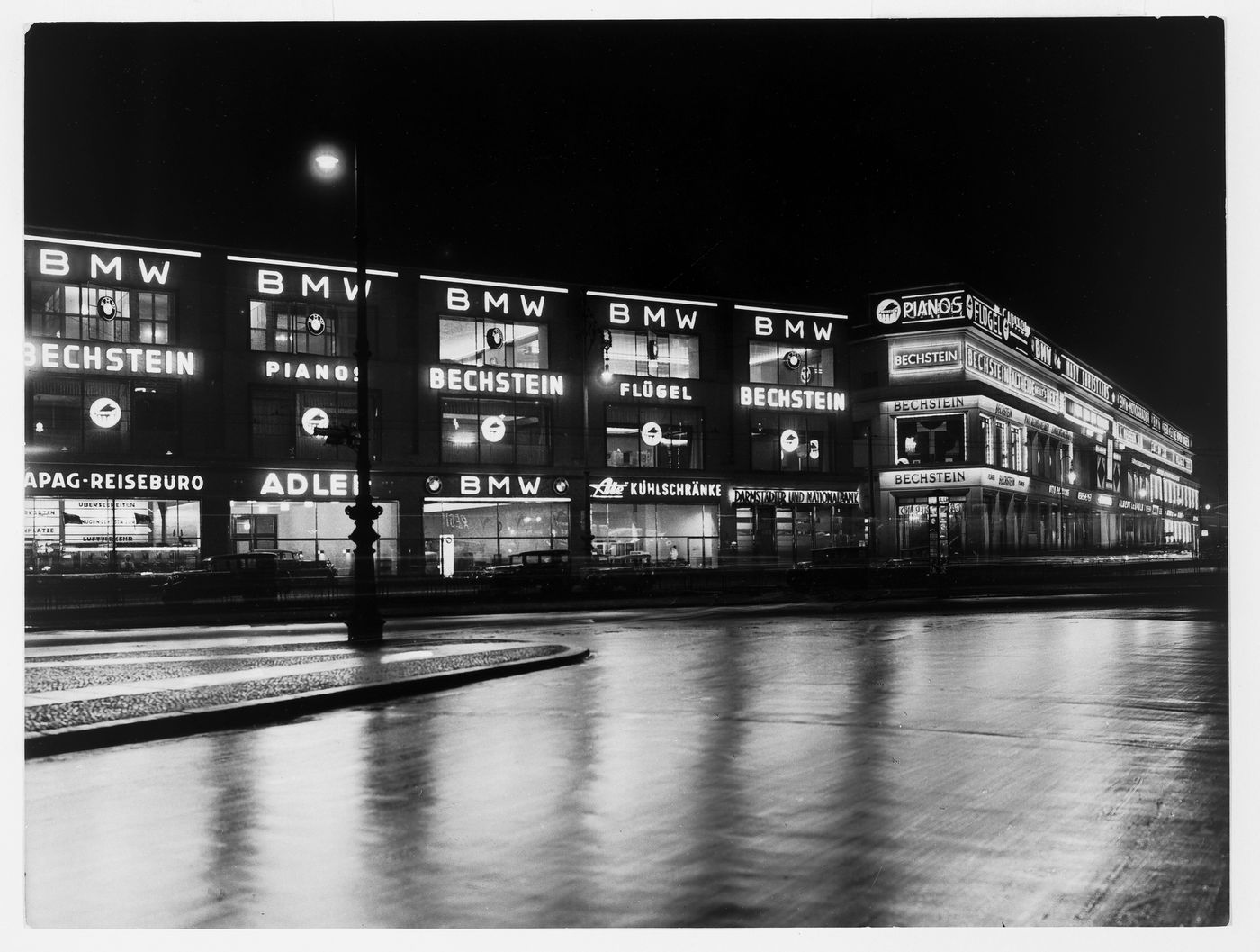 Night view of illuminated signs in long building (BMW, Bechstein, and others) seen from across street, Germany