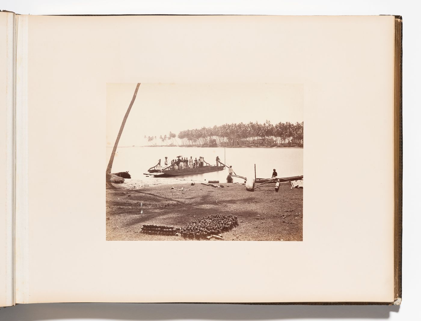View of a shore showing a boat and people with bottles in the foreground and trees and buildings in the background, Ceylon (now Sri Lanka)
