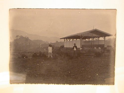 View of two men and a donkey in front of a barn [?] with hills in the distance
