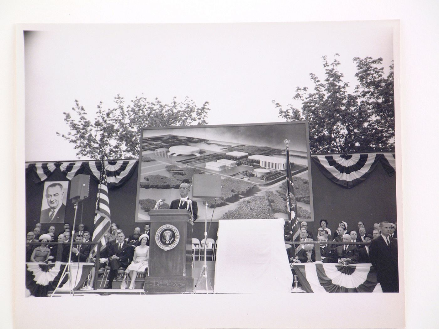 View of the dedication ceremony of the John F. Kennedy Educational, Civic and Cultural Center showing a partial view of the platform with people, posters of President Lyndon B. Johnson and a photograph of the model of the building, Mineola, New York