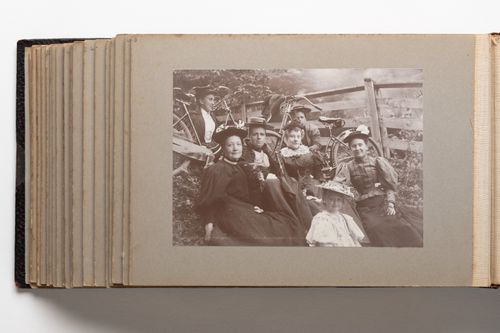 View of a group of six women and one young girl seated on the ground in front of a fence in a rural setting
