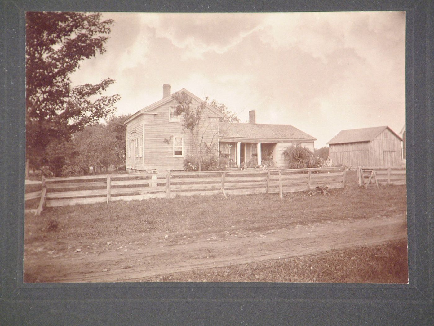 Wooden house with porch, barn adjacent, dirt track and wooden fence in foreground, New York State [?]