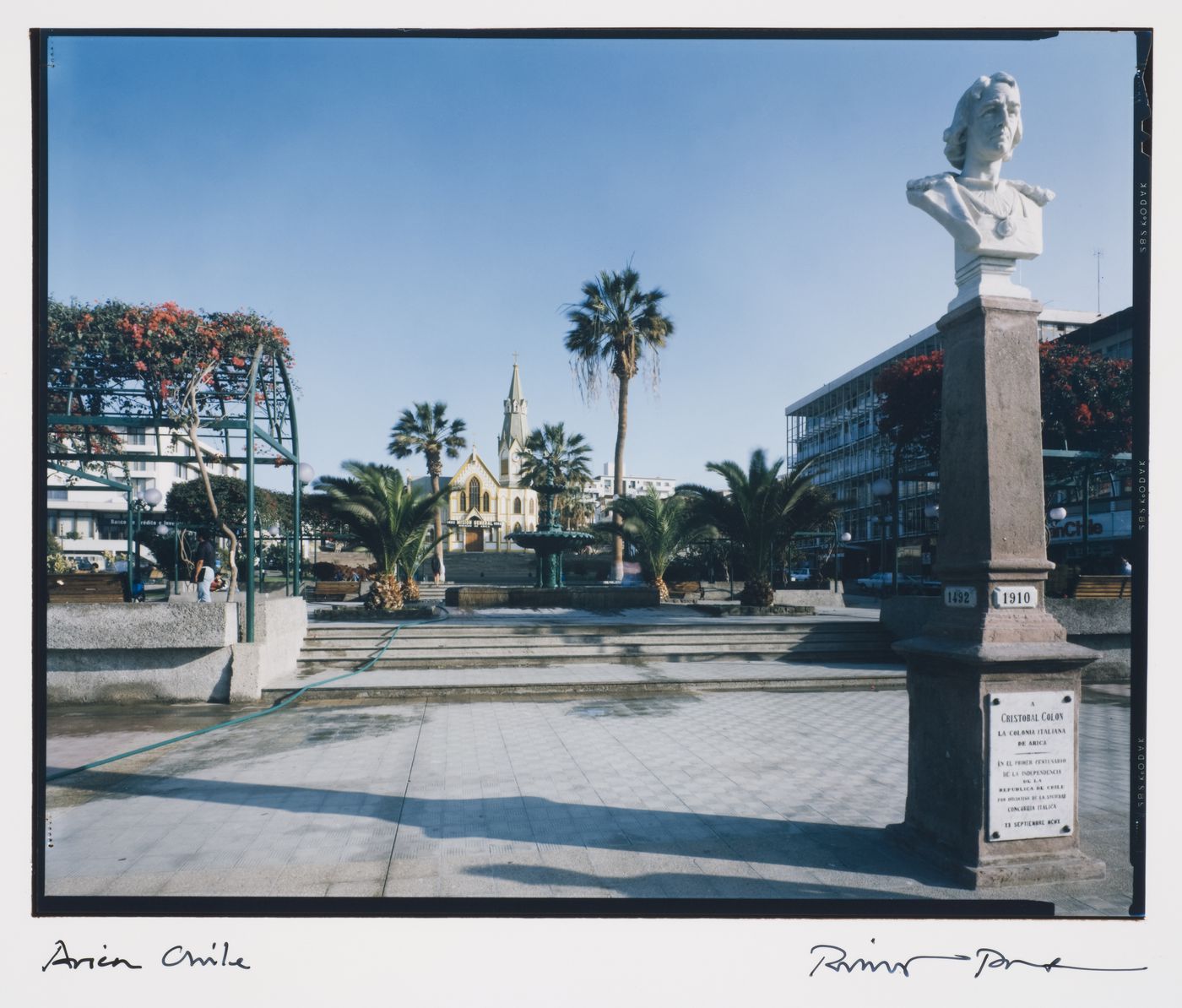 View across the Plaza Colón showing a fountain, trees, the principal façade of the Church of San Marcos and with a bust of Christopher Columbus [Cristóbal Colón] on top of a monument in the foreground, Arica, Chile