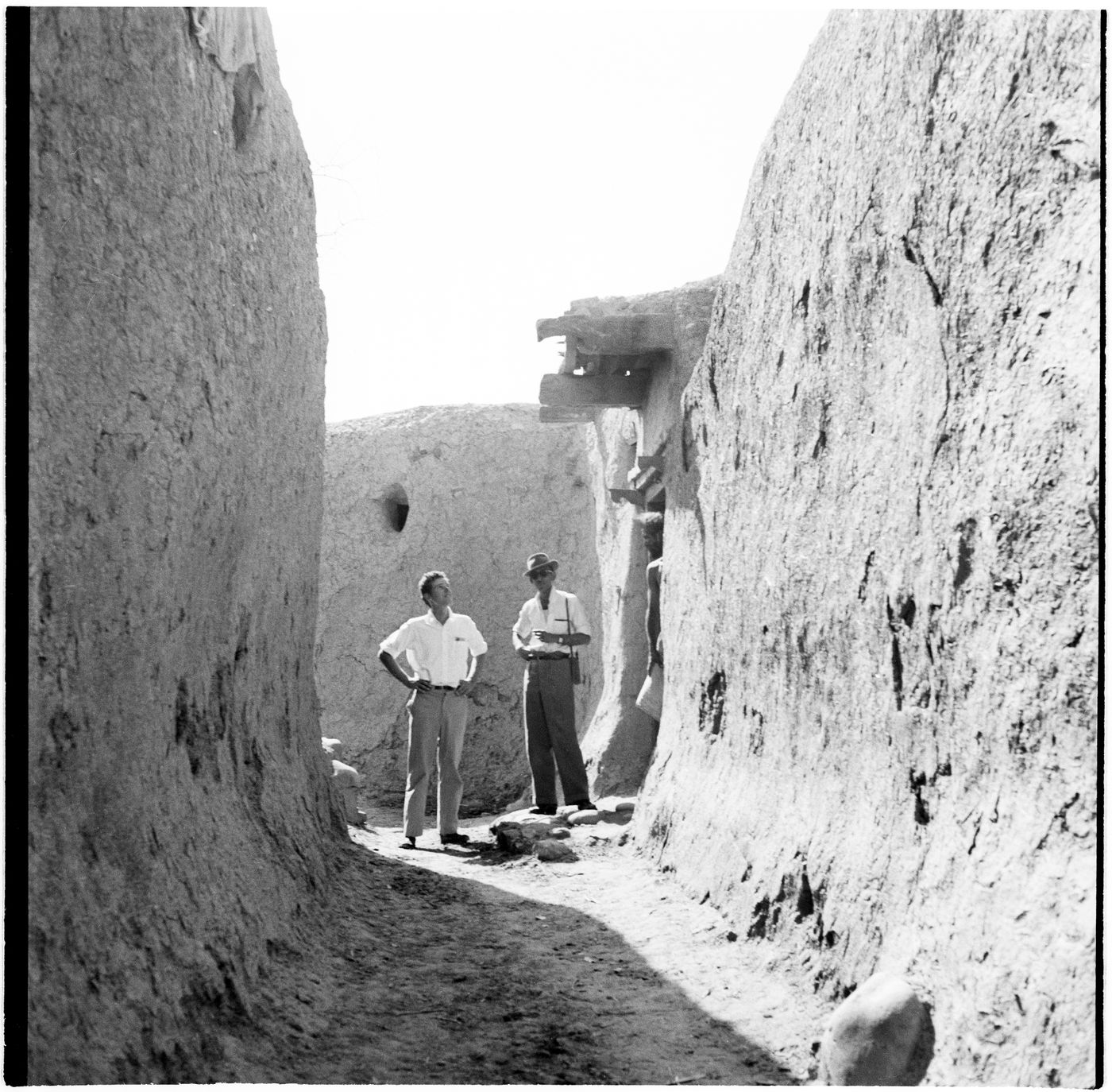 View of adobe structures in a village near Chandigarh, India, with 2 men standing in the centre and 1 man leaning in a doorway
