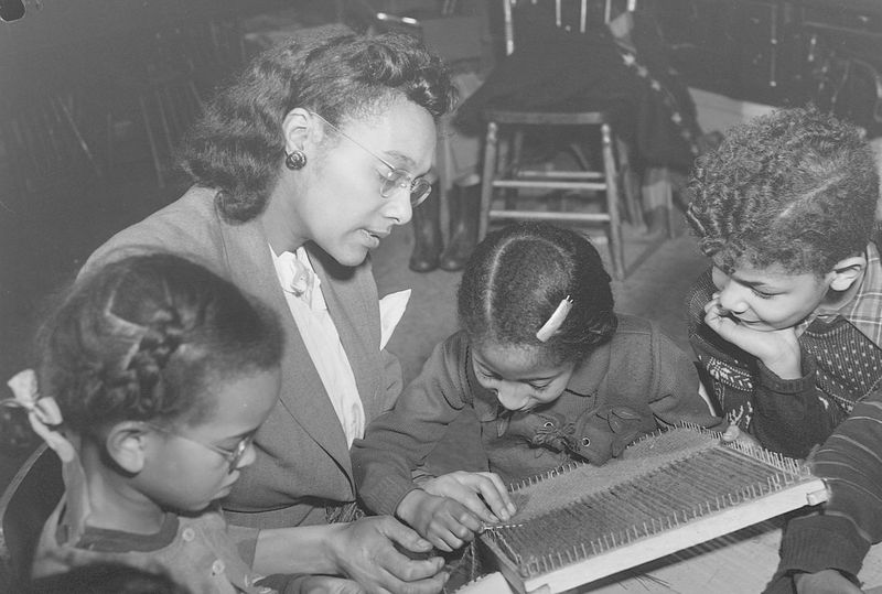 Des enfants, assis autour d'une table, tissent à l'aiguille avec l'aide d'une éducatrice